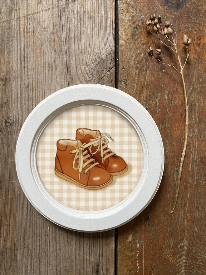 a photograph of a framed drawing of a little pair of brown baby boots, first walkers framed in an ivory wooden circular vintage frame hand drawn illustration drawn in a vintage story book style 