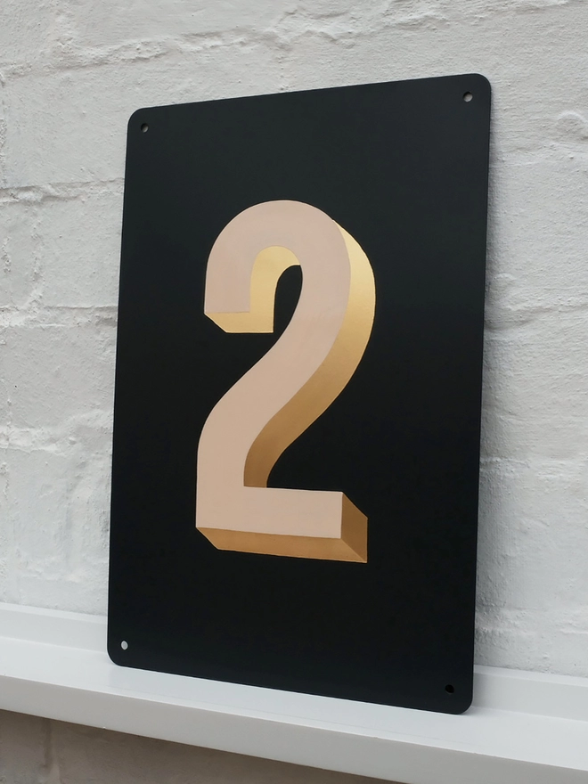 Peach and gold leaf house number 2, on anthracite grey metal plaque, against a white brick wall.