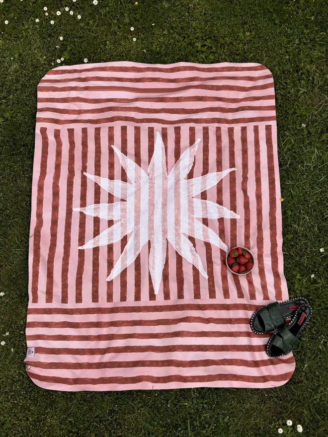 sun stripe fleece blanket. a burgundy and pink striped picnic blanket on grass.