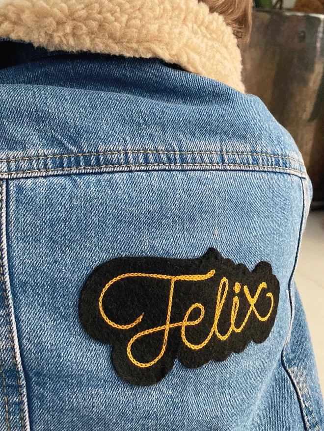 Black felt with gold embroidered lettering personalised patch reading the name 'Felix' attached to the back of a toddlers denim jacket