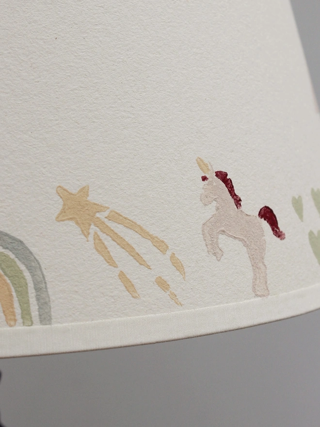 Close-up of a Studio Zef children's unicorn lampshade, featuring a hand-printed white unicorn with a burgundy mane and tail, a golden shooting star, pastel rainbow and pale green hearts. The whimsical illustrations are printed onto textured cream card in soft, muted colours, creating a charming, personalised lampshade design for a girl's bedroom or nursery.