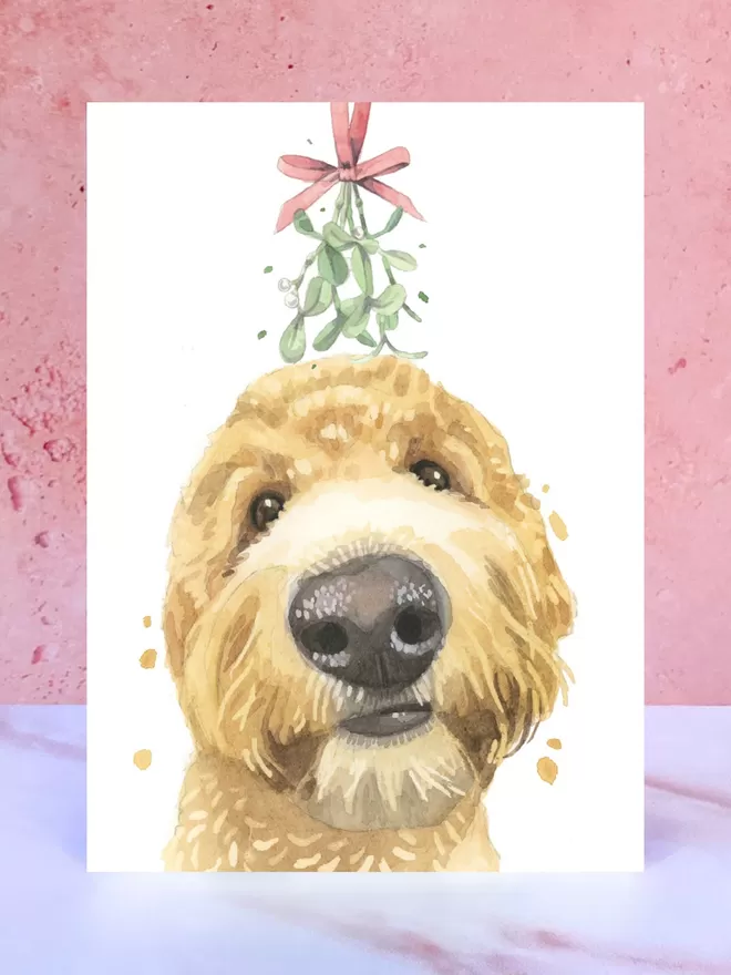 A Christmas card featuring a hand painted design of a Golden Doodle, stood upright on a marble surface.