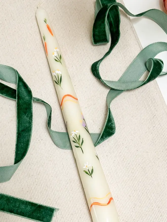 Carrots And Flowers Spring Candle, a spring candle lying on a plain surface accompanied by a green ribbon. It's floral design can be seen wrapping around it. 