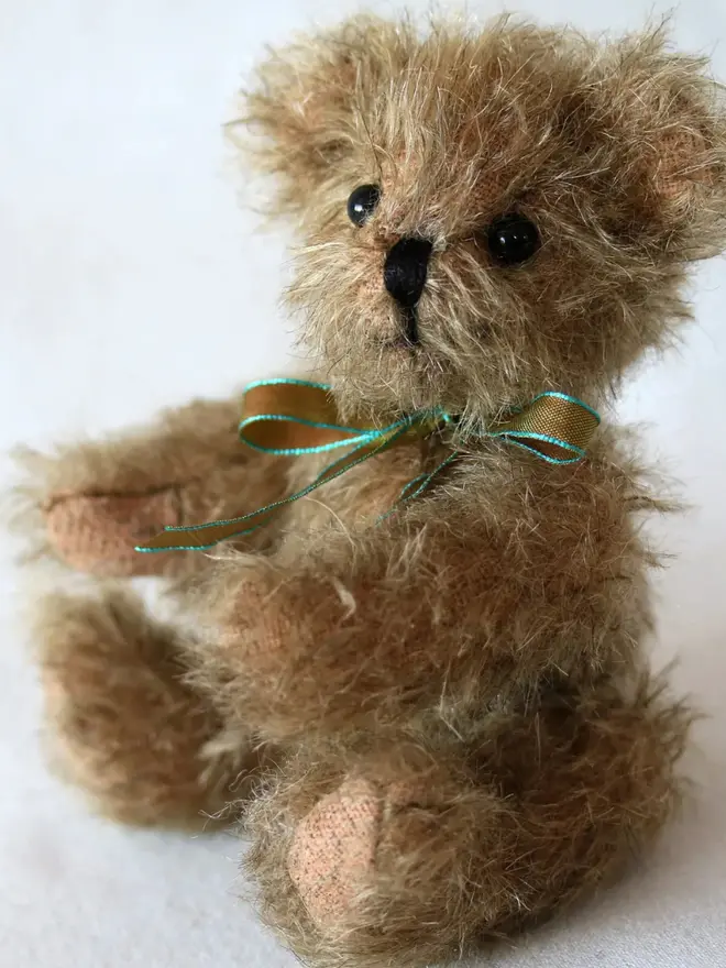 Small handmade brown teddy bear with a ribbon bow sitting against a plain background.