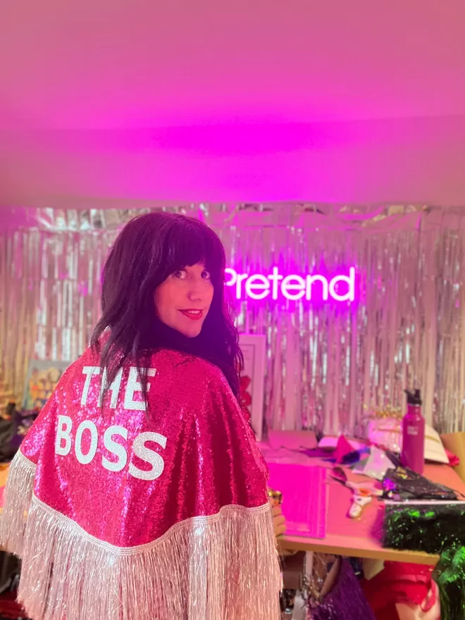 An adult woman wearing a midi cape with hot pink sequins, silver text reading 'THE BOSS' and silver tinsel.