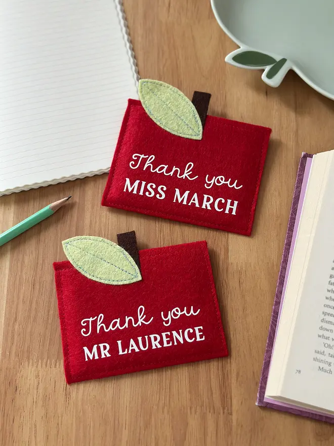 Two handmade red felt apple-shaped envelopes with green felt leaves and brown stems, each reading "Thank you MR LAURENCE" and "Thank you MISS MARCH", displayed on a wooden desk with a pencil, notebook, and book. It's such a sentimental personalised, handmade teacher thank you gift.