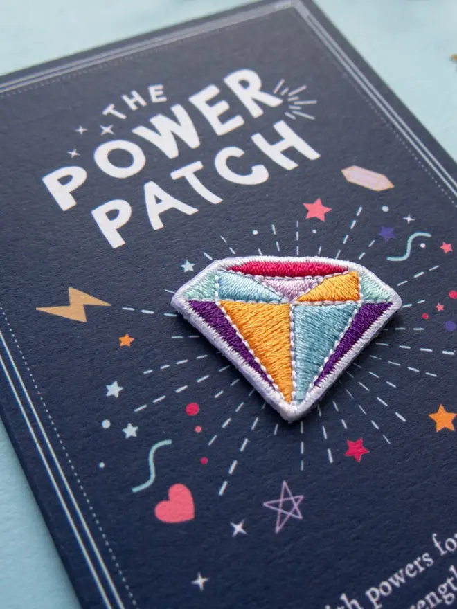 Close-up of an embroidered gem patch in orange, turquoise, purple and pink, mounted on a navy backing card. White "The Power Patch" lettering, stars and hearts surround it.