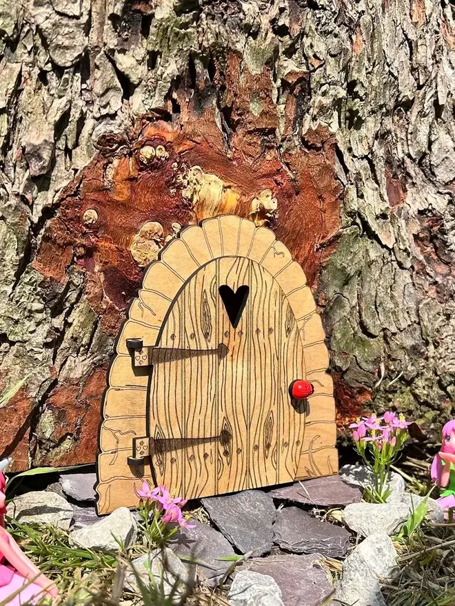 Wooden fairy door for kids with engraved woodgrain detail, heart-shaped window, and decorative hinges, displayed at the base of a tree surrounded by moss and wildflowers.