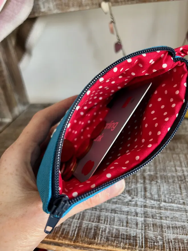 Hand holding open a blue coin purse with zip opening and red polka dot lining, holding bank card s.