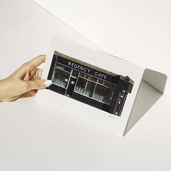 A hand with blue nail polish holding a print featuring an illustration of the Regency Cafe’s black tiled shopfront against a white background.