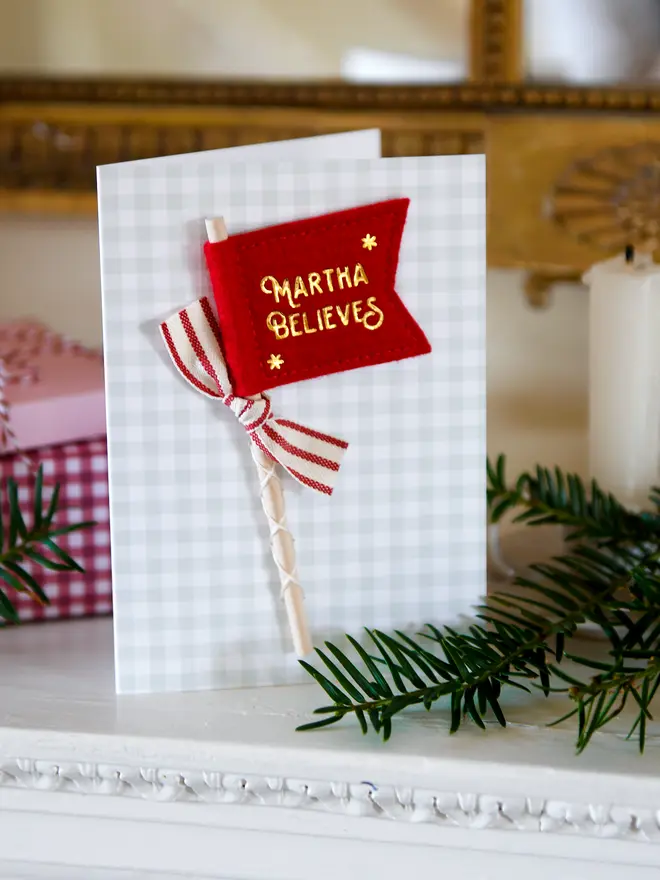 A Christmas card with a tiny handmade felt flag attached. The flag reads 'Martha Believes' and has a red striped ribbon tied to it.