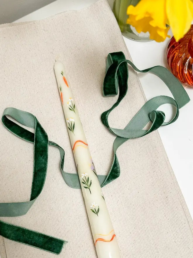 Carrots And Flowers Spring Candle, a spring candle lying on a plain surface accompanied by a green ribbon. It's floral design can be seen wrapping around it. 