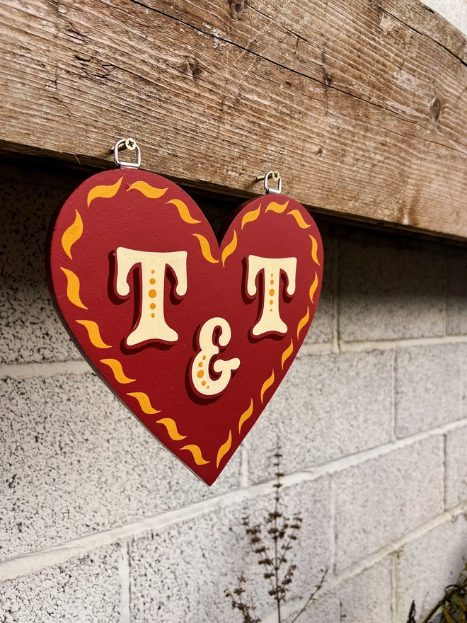 Wooden love heart cut out with a red matte background, glossy golden yellow curve details painted along the edges and ivory lettering with dark red shade. It is hanging on a wooden planter.