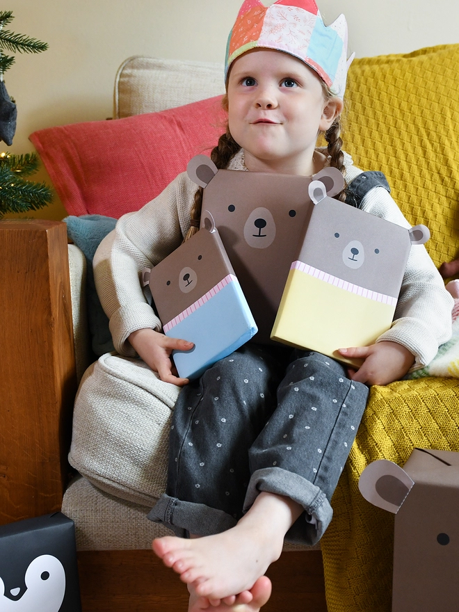 A young girl sitting on a sofa beside a Christmas Tree is holding three gifts wrapped up as brown bears.