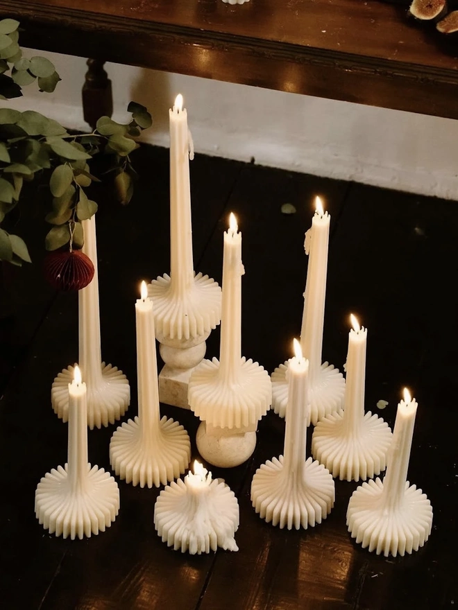 Pair Of White Flair Candles (Set Of 2). A top-down view of lit cream fluted candles grouped on a dark floor around a stacked stone holder, with eucalyptus and a red paper ornament. The warm glow of the collection creates an inviting, atmospheric display.