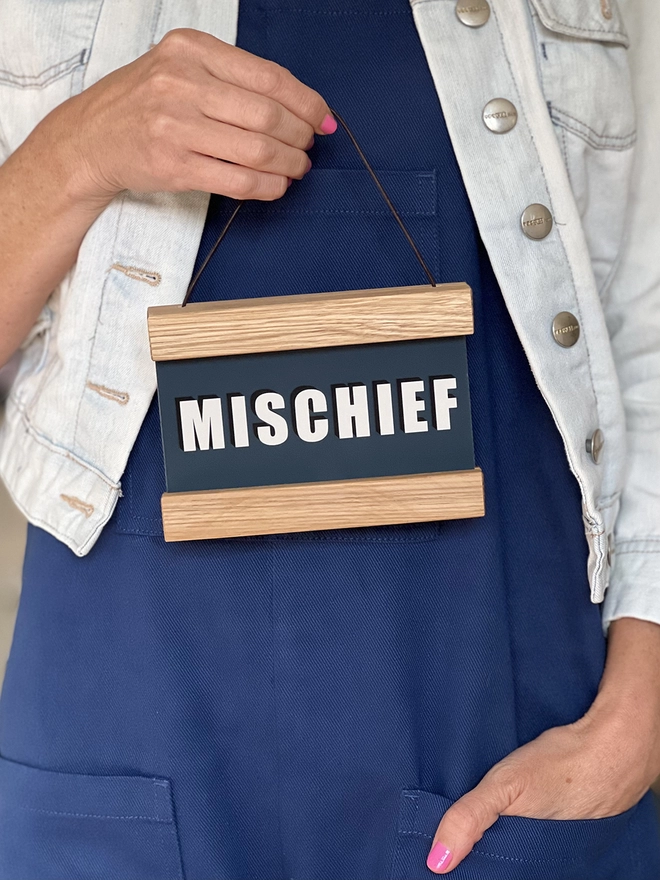 A print in a Navy colour with the word, MISCHIEF framed in a oak hanger, with a waxed cotton cord for hanging being held by a hand with pink nail varnish 