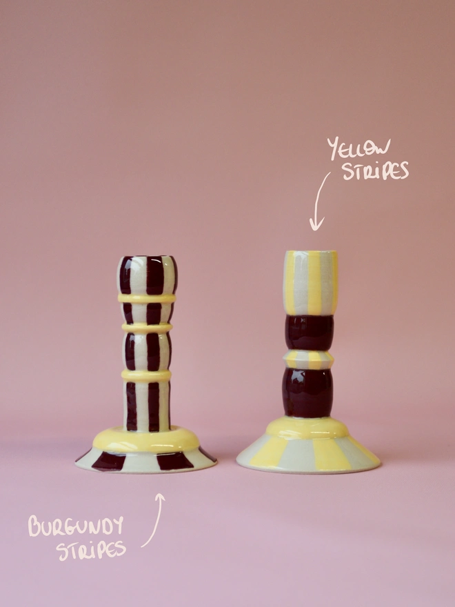 Handmade ceramic candlesticks with burgundy and yellow stripe patterns, including a tall burgundy striped design and a shorter yellow striped candlestick, set against a pink background.