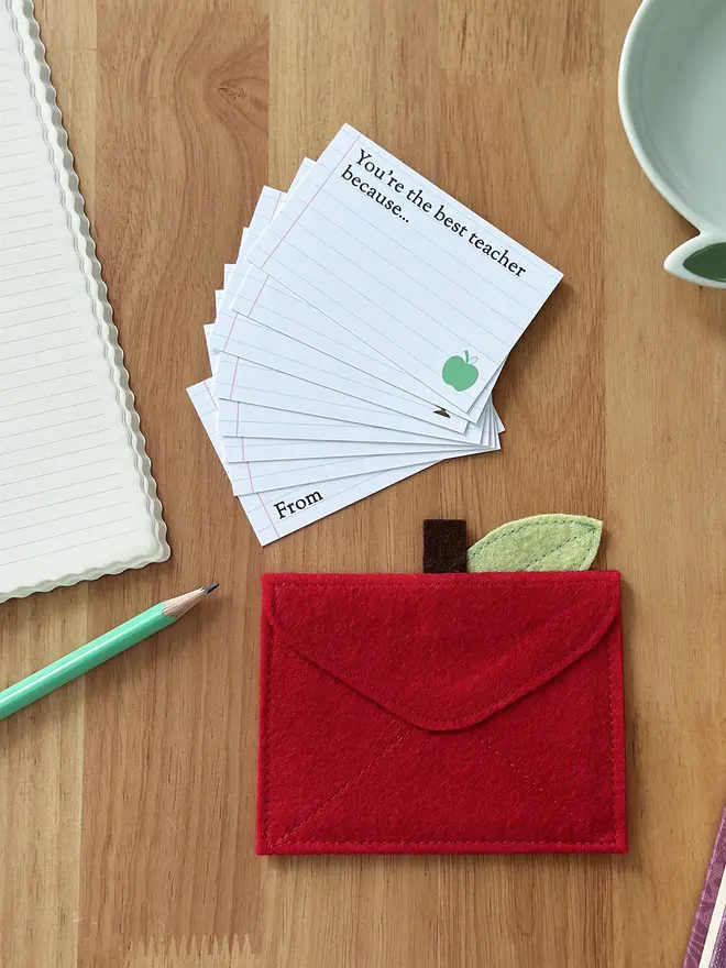 A handmade red felt apple-shaped envelope lays upside down. The heirloom apple envelope is surrounded by prompt cards that can be completed by a child, displayed on a wooden desk. It's a thoughtful gift for teachers from children.