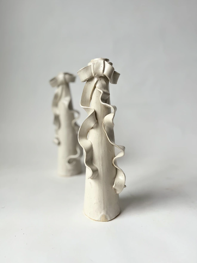 two cream white stoneware tall candlestick holders with a ceramic bow with long ribbons flowing down the body of the candlestick itself