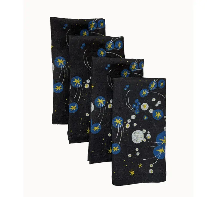 Solaris Napkins In White Or Blue. Set of four blue cloth napkins with blue and yellow starburst and orbital patterns, stacked in a staggered arrangement.