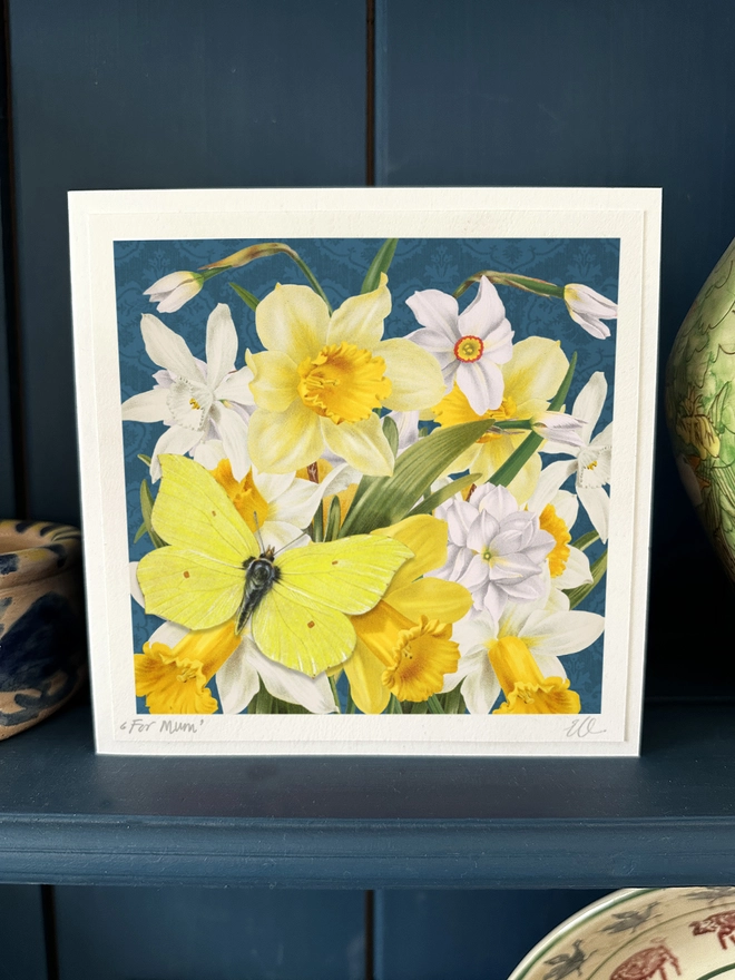 Mother’s day card, showing beautiful daffodils and white narcissi with a white butterfly. The card can be personalised on the bottom left corner. The camellias are against a stylish grey background. Sitting on the flower is a yellow Brimstone butterfly. The butterfly is 3 dimensional and the wings can be folded open for display. The card is sitting on a dresser displayed in the home setting.