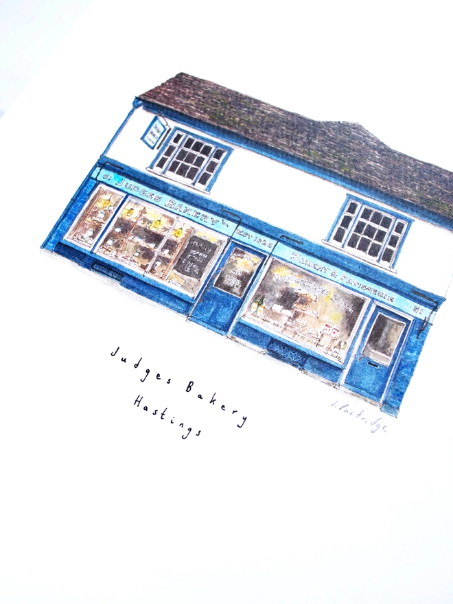 Watercolour illustration of Judges Bakery a beautiful blue shopfront with windows full of bakery goods sits below a white first floor and dark brown / grey roof.  The photo is at an angle showing some of the intricate detail of the painting. 