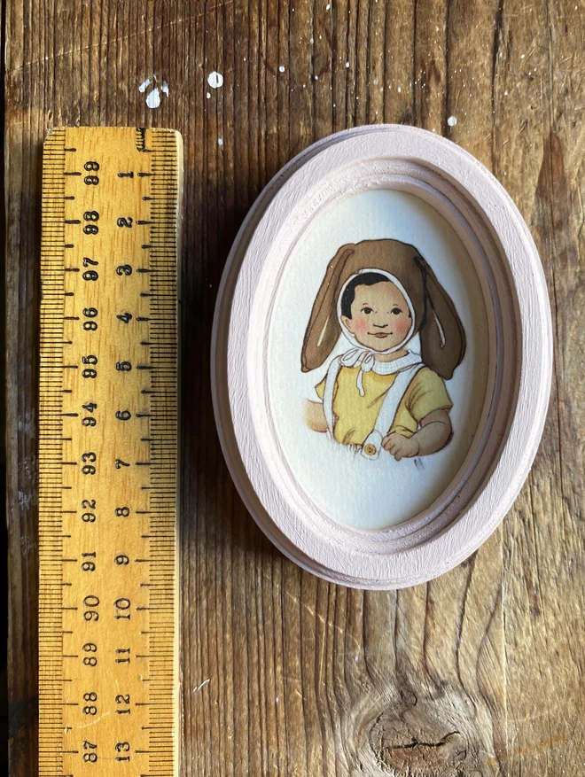 a photograph of a little boy drawn in a childrens vinateg book style wearing an easter bunny bonnet faremd in a dusty pink wooden oval framed photographed next to a vintage ruler