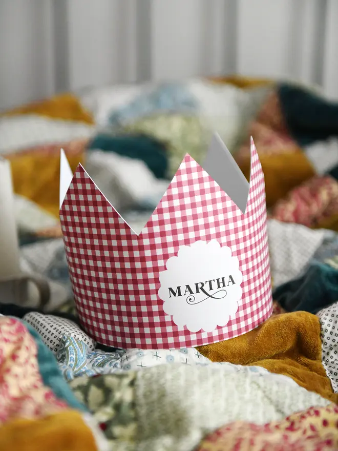 A red gingham Christmas crown, with a scalloped circle on the front and the name Martha, rests on a cosy patchwork quilt. 