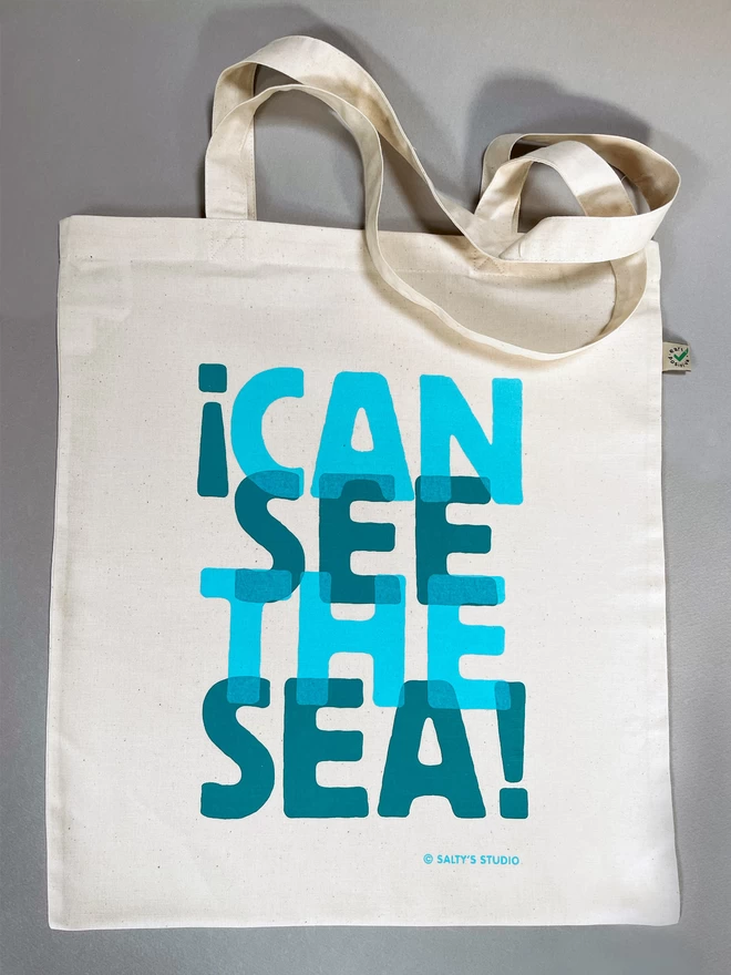  The words I Can See the Sea is screenprinted in two overlapping turquoise blues on this fairtrade cotton tote bag. Flat laid on a grey background, with the handles soft folded at the top.