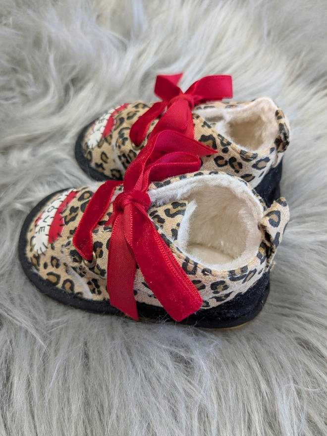 A pair of leopard print baby booties, lined with fur and with red velvet ribbon laces. A red heart with a white scroll adorns each booty. One says BABY, the other, LOVE in black stitches