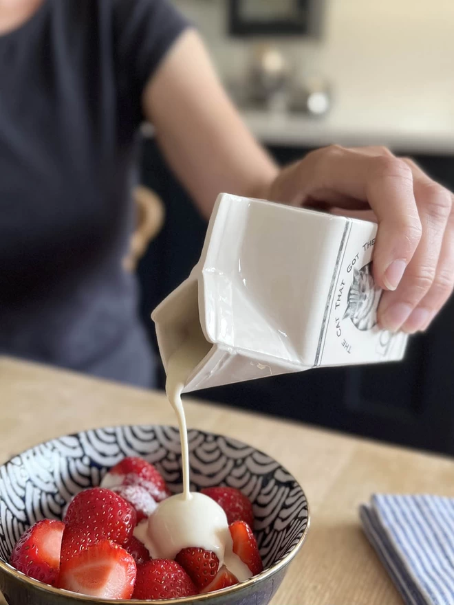 A small handmade ceramic carton is pouring cream over a bowl of fresh strawberries.
