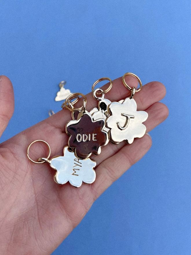 a hand holds finished brass pet tags. they are flower shaped and with a shiny finish with dogs names engraved onto the fronts.