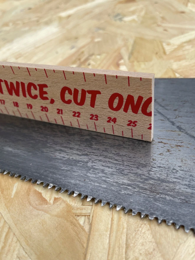 Part of a wooden ruler in shot, with graphic lettering printed on it, ‘Measure Twice, Cut Once’ sat on a saw, with a chip board background.