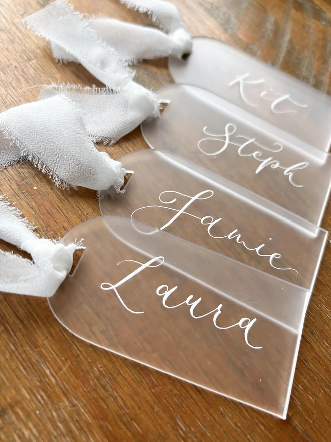 Personalised Calligraphy Frosted Acrylic Place Cards, frosted acrylic place cards with white chiffon ribbons grouped together on a wooden table. 