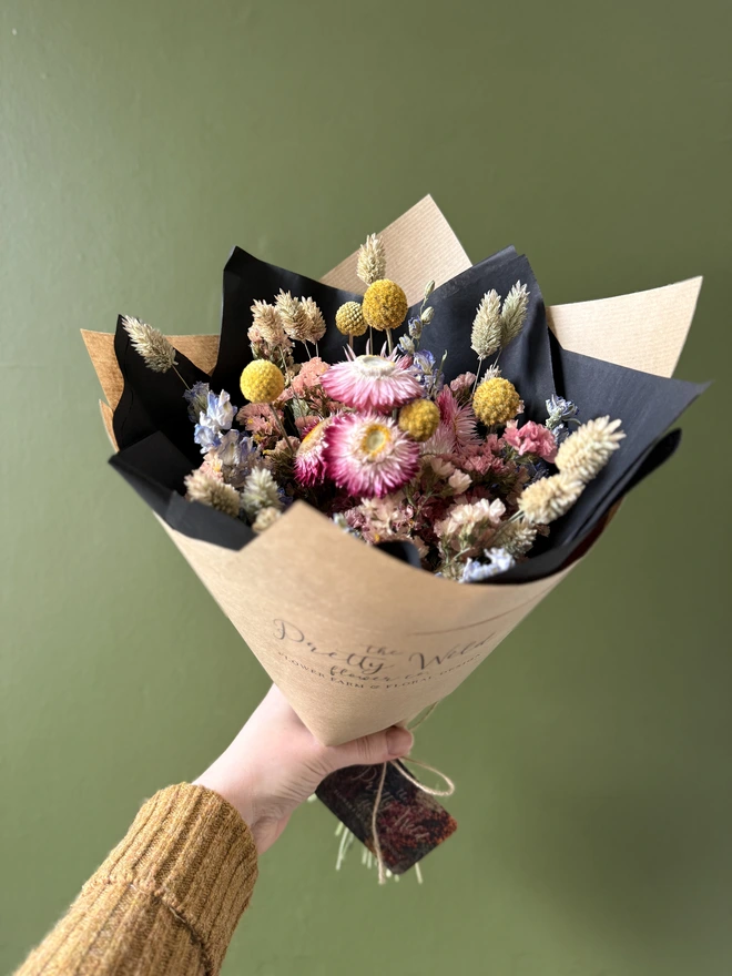 a bouquet of dried flowers in bright colours wrapped in black tissue and kraft paper held by a person in a mustard coloured jumper with their left arm against a green wall