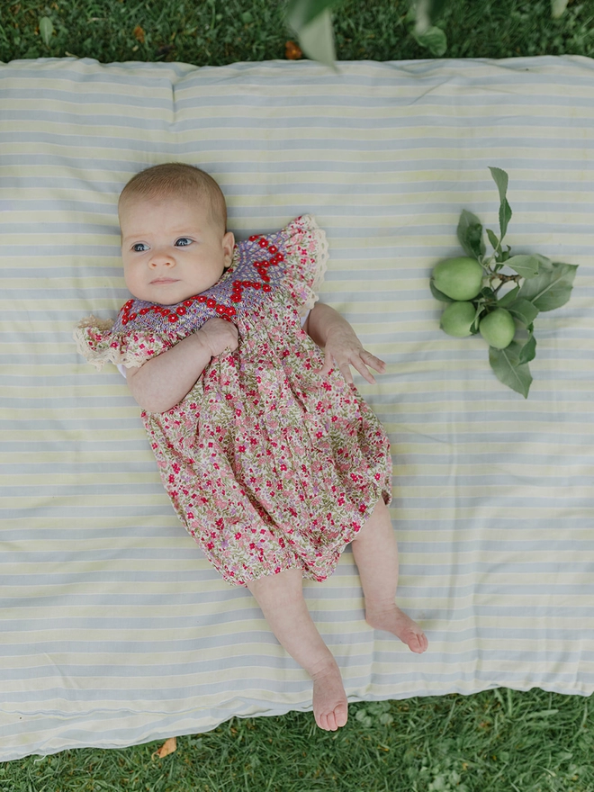 A baby in a red floral romper with bishops smocking lies on a striped mattress on the grass