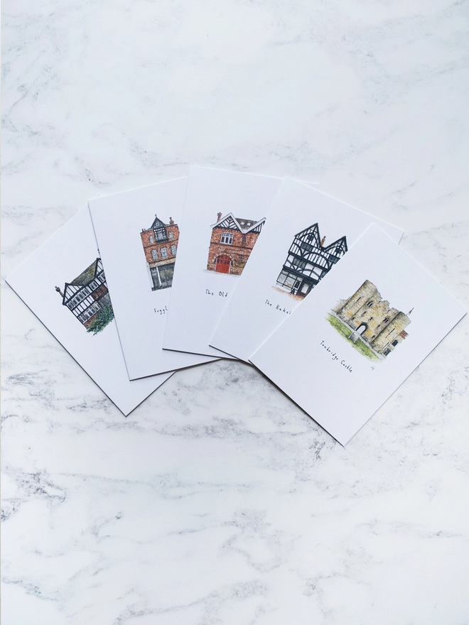 A photo of a set of five greetings cards featuring buildings / small businesses in Tonbridge, Kent. The set includes 65mm Coffee, The Old Fire Station, The Bakehouse at 124, Fuggles Beer Cafe and Tonbridge Castle. These are a photo of the set of A6 greetings cards to illustrate the different designs. The cards sit fanned out on a white marble backdrop. 