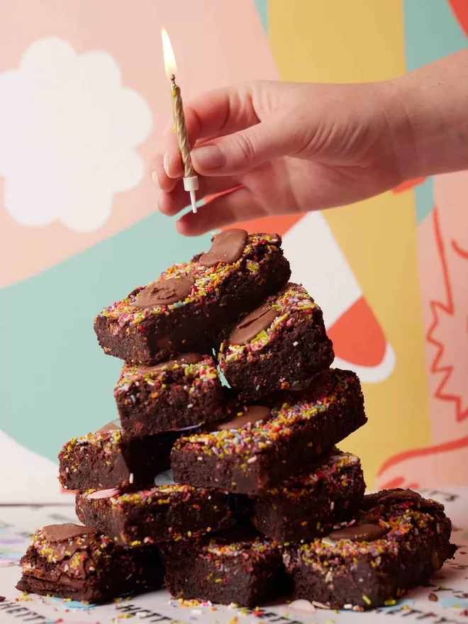 Funfetti brownies with sprinkles and milk chocolate buttons stacked for a Birthday celebration with a hand adding a lit candle to the top