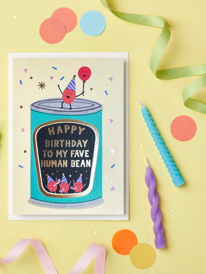 Happy Birthday To My Fave Human Bean card paired with a white envelope on a yellow background with confetti and streamers scattered around.
