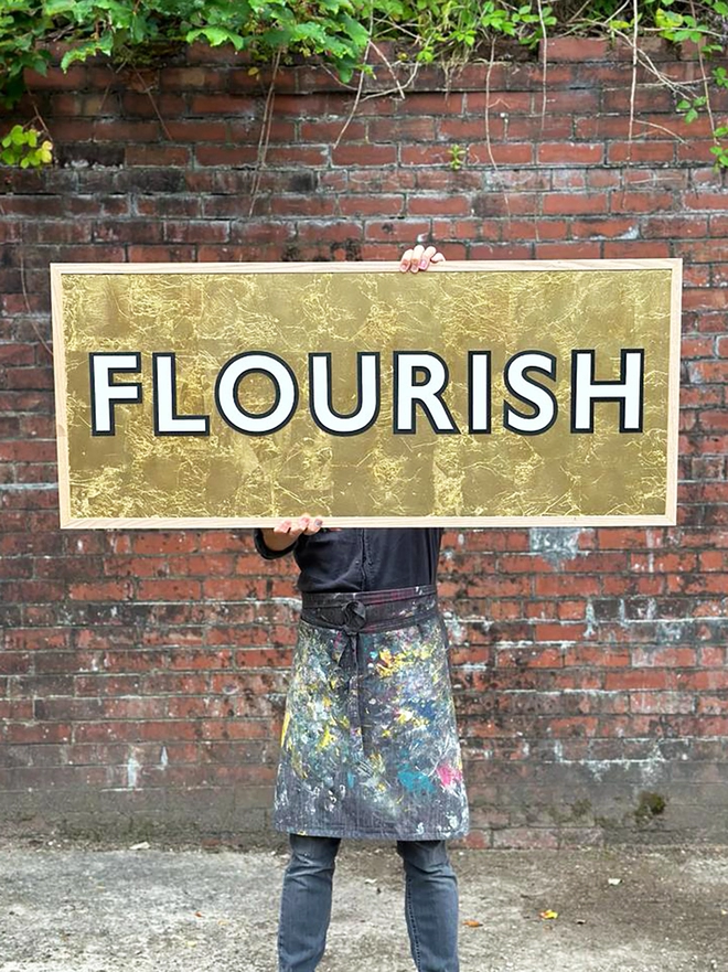 A person wearing a painted splattered apron holding a gold sign reading FLOURISH at head height 