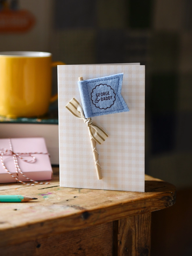 Personalised Father’s Day card with soft blue felt flag reading “George & Daddy”, handmade keepsake card with ribbon and wooden stick detail. The card stands on a wooden table beside a book, mug, candle, and wrapped gift.