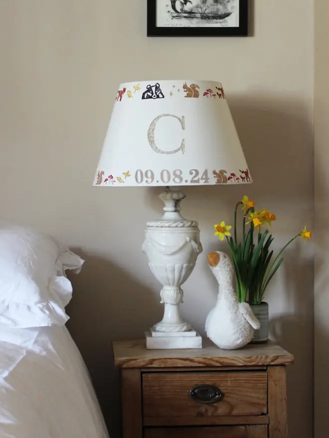 A cosy children's bedroom with a rustic wooden nightstand featuring a personalised lampshade with the letter "C" and a countryside pattern. A plush goose, daffodils, and white bedding add charm. Neutral empire light shade with pretty animal details, ideal for kids bedroom design and personalised kids gift.