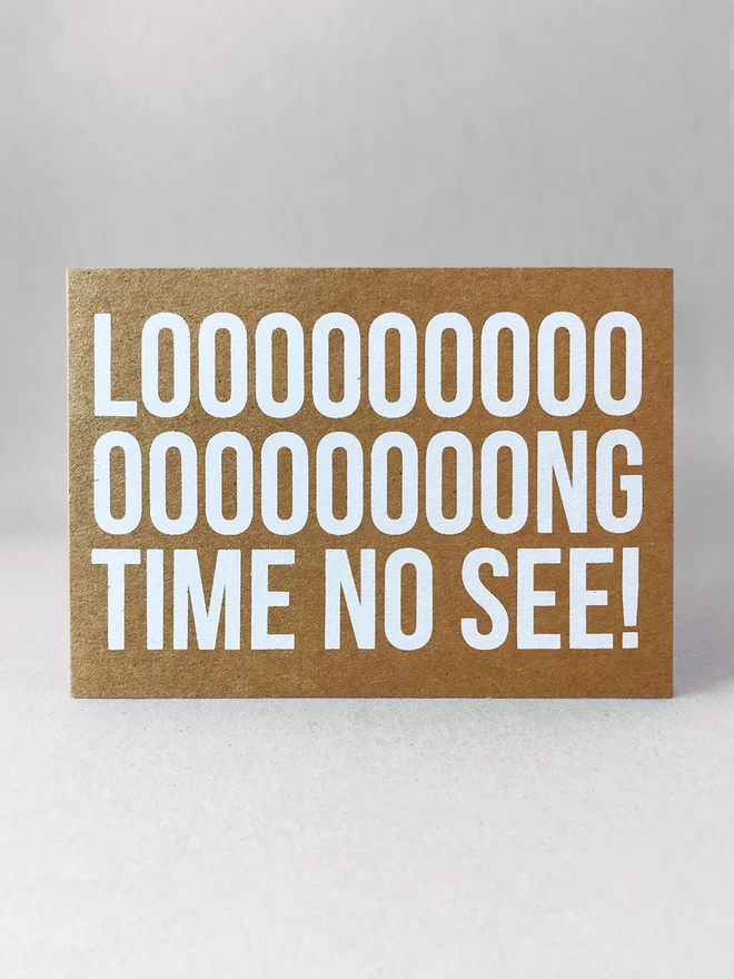 Looooooong time no see screenprinted across this card with multiple o’s in the Long, emphasising the word LONG. Printed in white ink on a brown kraft card stock in landscape format. Stood front on, in a light grey studio set.