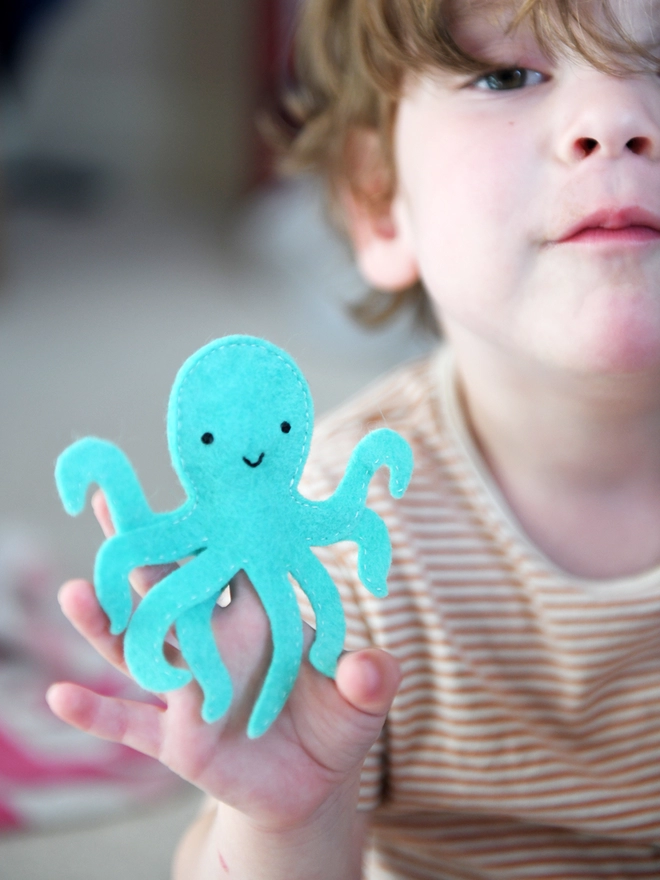 A young child is holding a turquoise felt octopus finger puppet on their fingers.