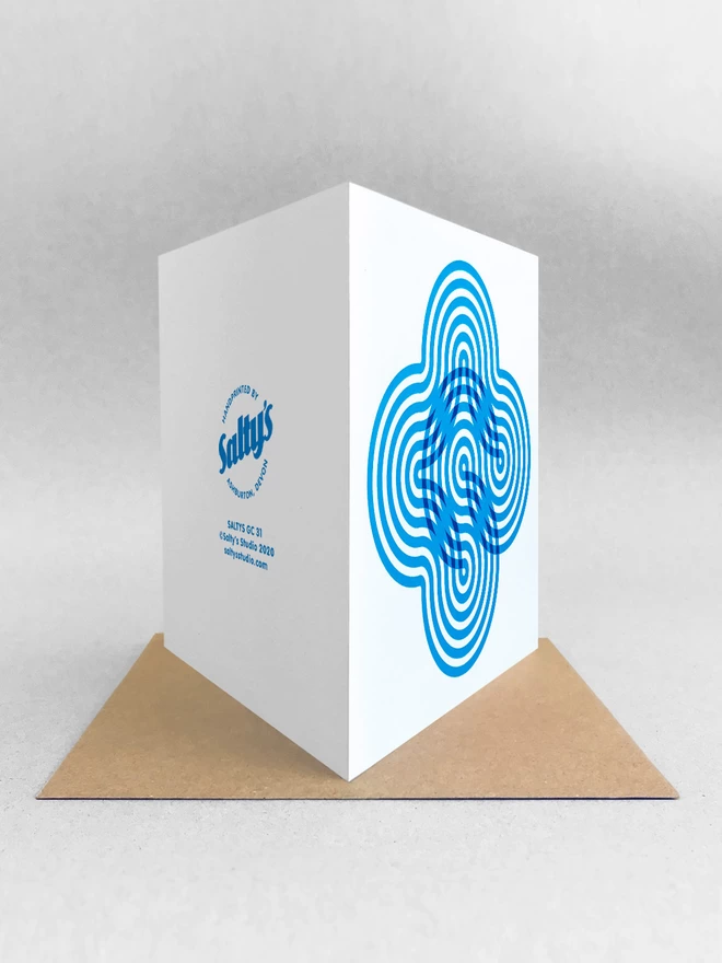 With its fold towards us we see the front and back of this square card. Showing the swirling blue stripes creating a flower-like visual on the front of this card and the logo and blurb on the back. This studio shot shows the card stood on a beige kraft envelope on light grey background.