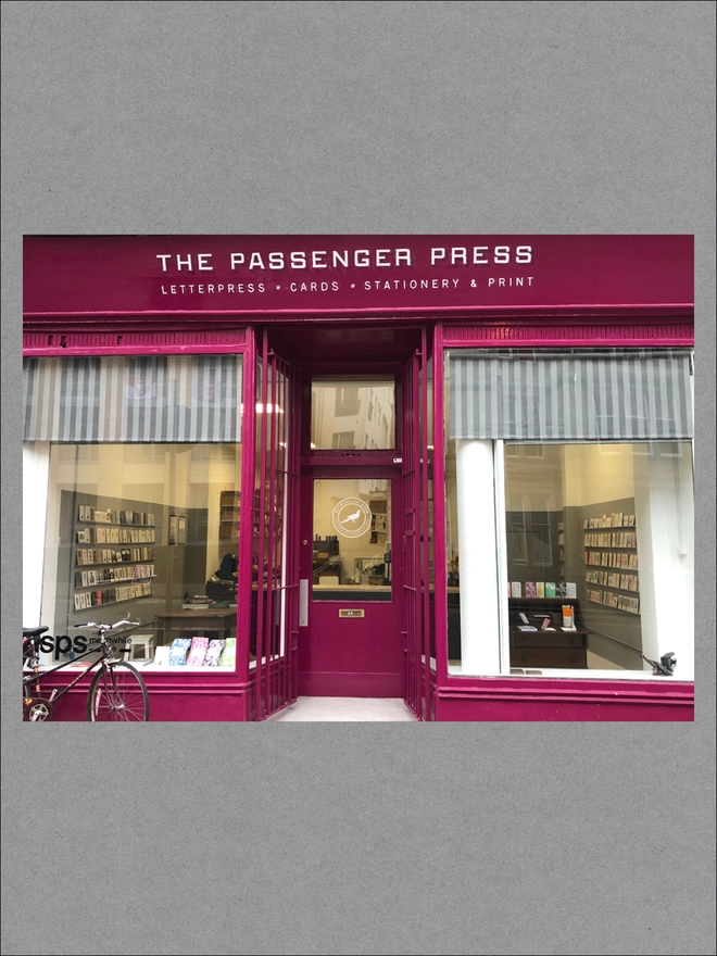 A Raspberry pink shop with striped curtains and a white sign reading 'The Passenger Press'.