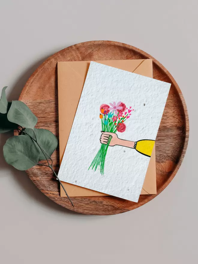 Seeded Paper Greeting Card with an illustration of a hand holding out a bouquet of flowers on a wooden tray next to a Eucalyptus branch