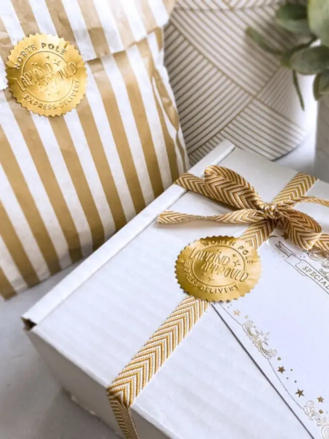A gold seal fastens a gold-and-white striped paper gift bag. Another seal holds a chevron ribbon in place on a white gift box.
