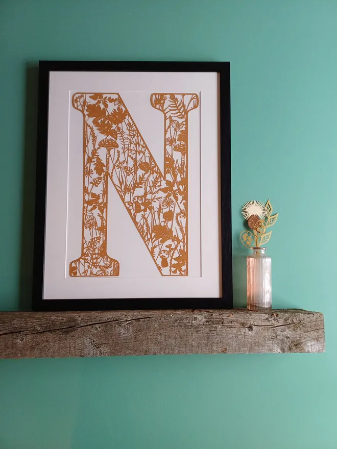 Papercut Letter N in Raw Sienna in Black Narrow Frame with off white mount displayed on wooden shelf against a green wall