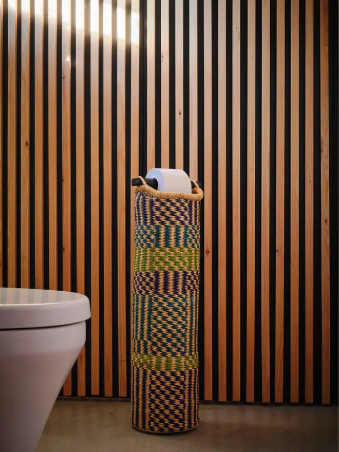 Ghanaian Hand Woven Tubular Toilet Roll Holder, a toilet roll holder sitting in a bathroom. It sits in front of a wooden backdrop. 